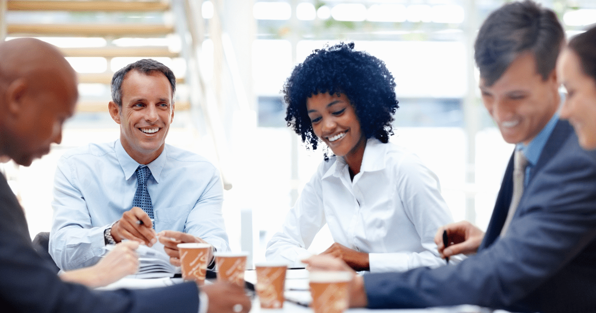 3 men and 2 women of different ethnicities are wearing smart business attire and sat around a boardroom table with paper and coffee cups on.